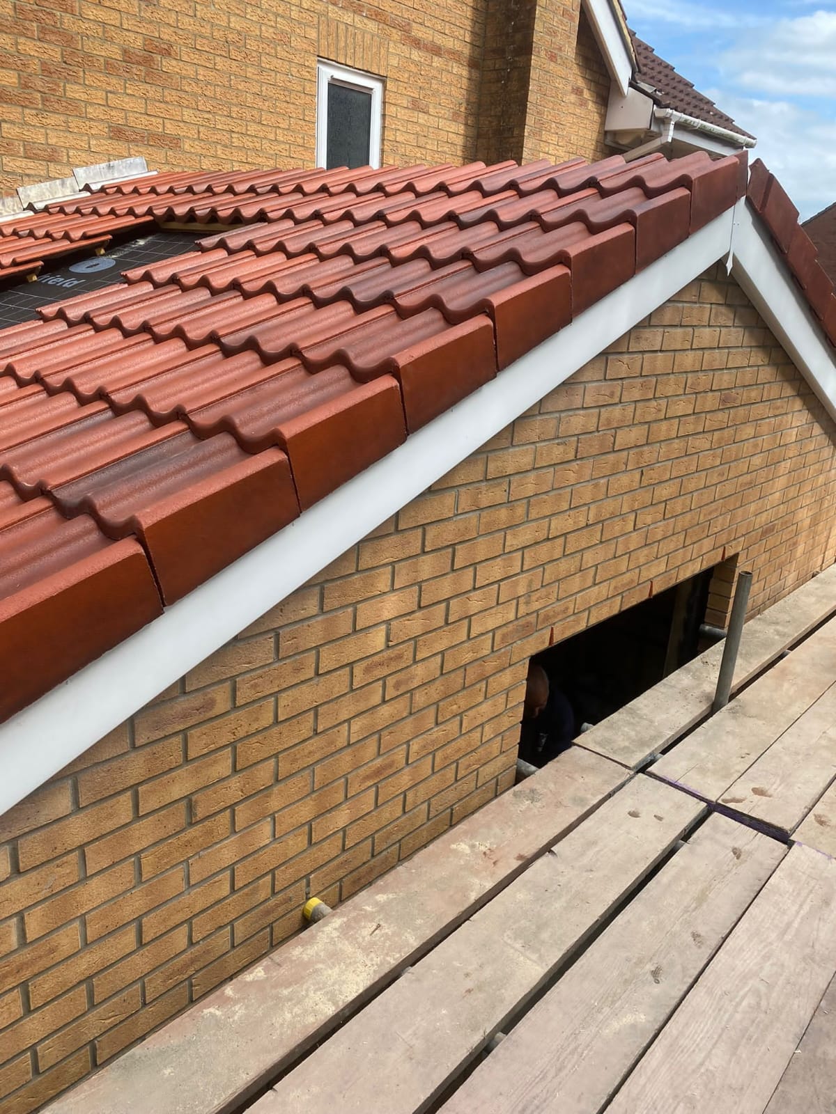A red-tiled roof with white trim on a brick house, viewed from a wooden scaffold platform. There's a small rectangular opening in the brick wall below the roof. The sky is partially visible.