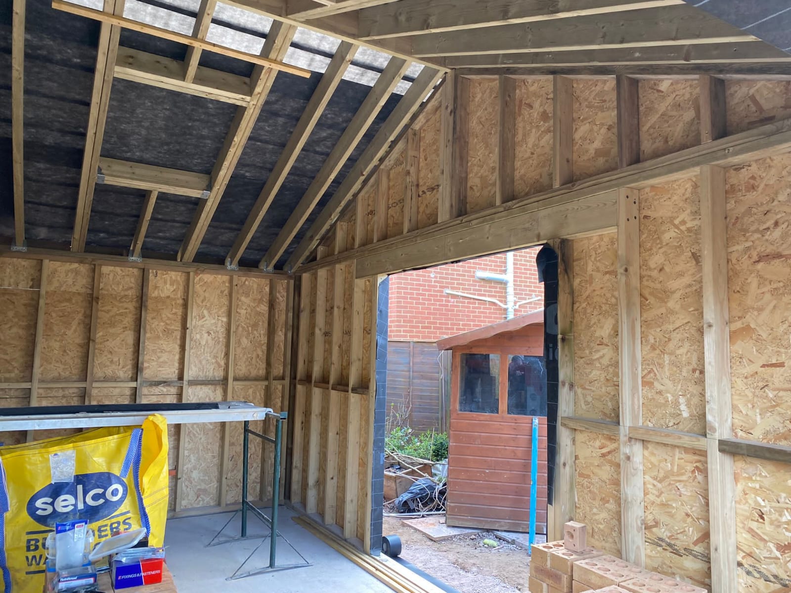 A construction site with wooden framework inside a partially built structure. A yellow Selco Builders bag is on the floor. In the background, there's a garden shed and some stacked bricks.