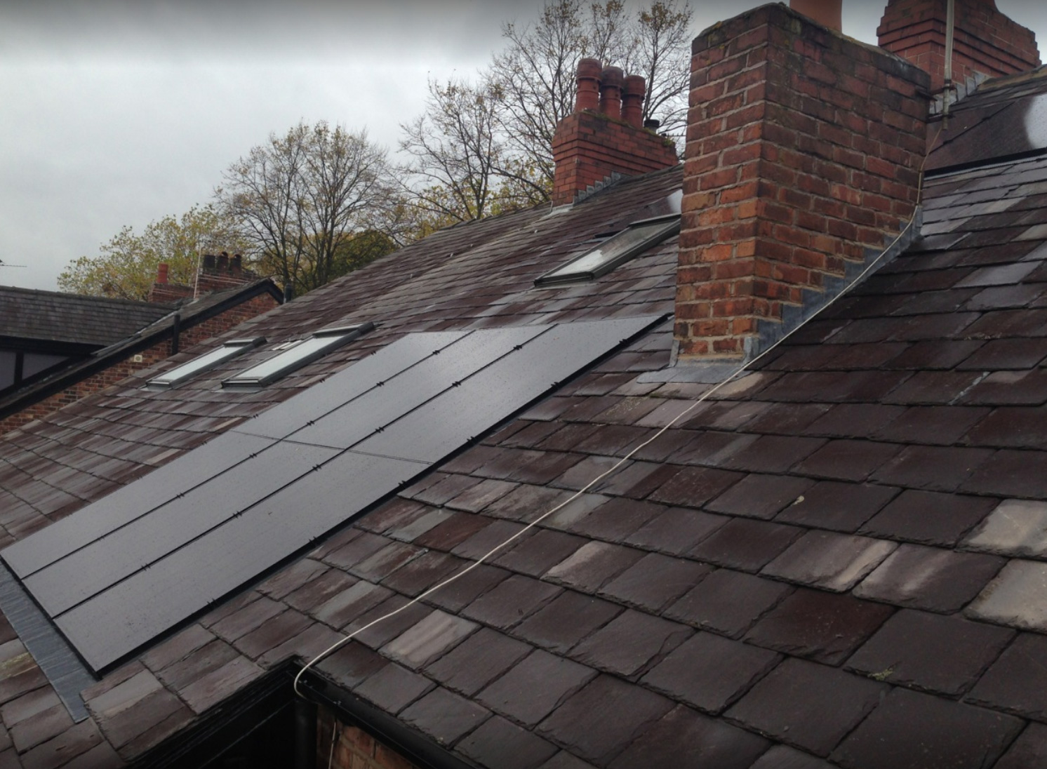 A slate-tiled roof with solar panels installed on it, set against a cloudy sky. The roof has brick chimneys and the surrounding trees show signs of early spring with sparse leaves.