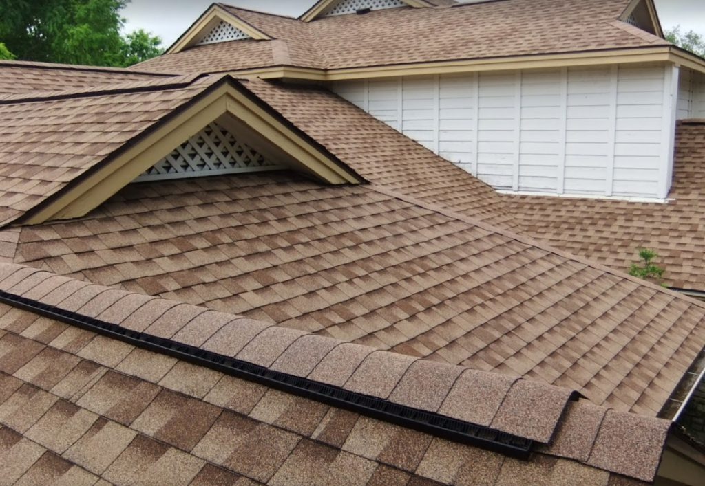 The image shows a close-up view of a house roof covered with brown asphalt shingles. The roof has multiple sections with triangular and rectangular shapes, and the shingles are aligned in neat rows. There are small trees in the background.