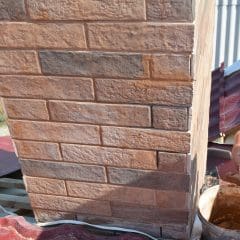 A person is repairing a brick chimney on a red corrugated metal roof. They are applying mortar to fill gaps between the bricks. A nearby container holds more mortar. Sunlight casts shadows on the roof and chimney.