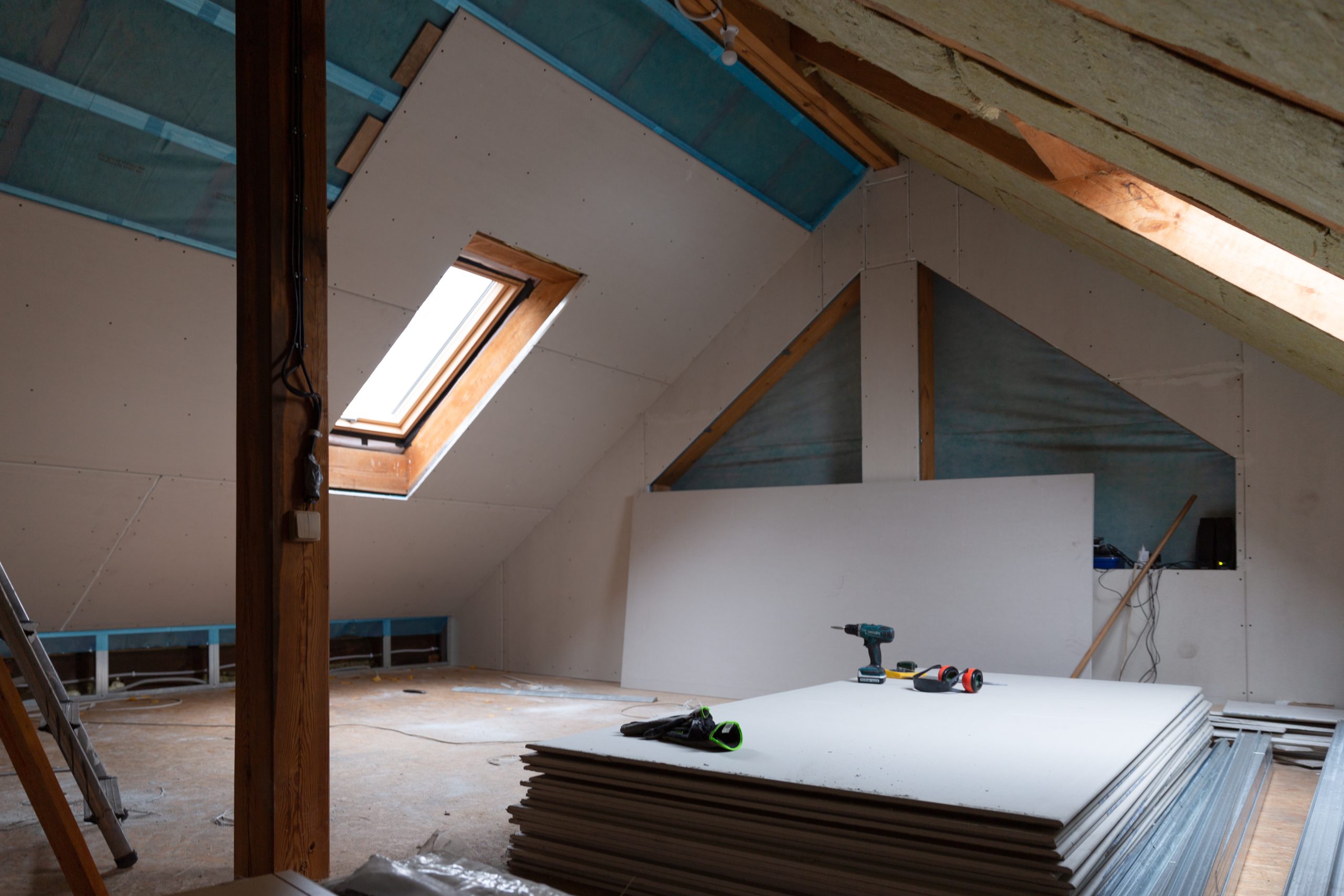 An unfinished attic renovation with drywall on the walls and ceiling. A window in the sloped ceiling lets in natural light. Tools and stacked drywall sheets are on the floor, with wooden beams and insulation visible.