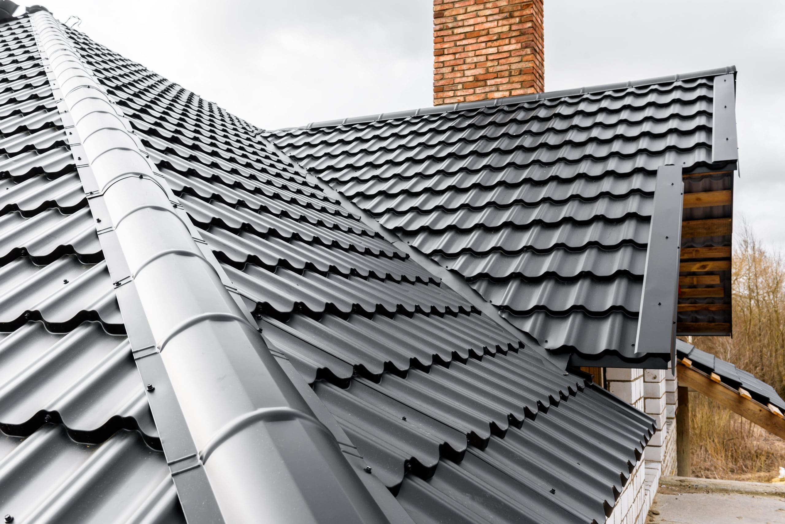 A close-up of a house with a modern black metal roof featuring a wavy pattern. A red brick chimney is visible, and the sky is overcast.
