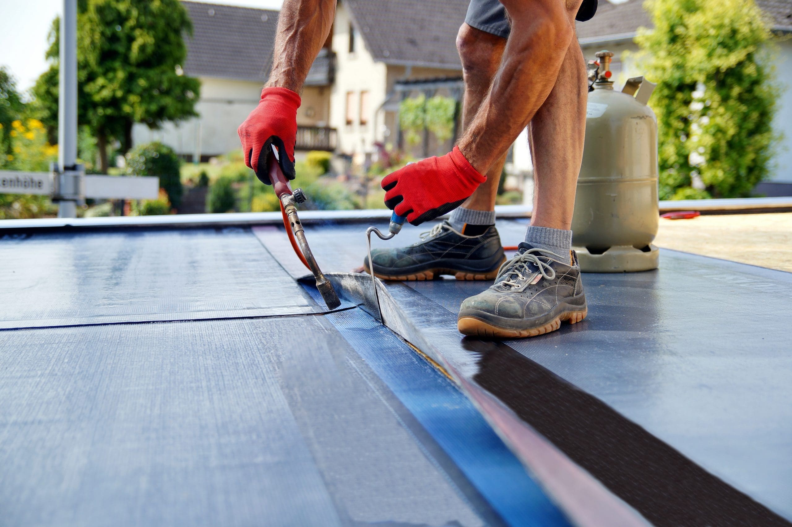 A person wearing red gloves applies a blowtorch to the edge of a flat roof, sealing a strip of material. Nearby is a gas tank. The scene suggests roof repair or maintenance in a suburban neighborhood.