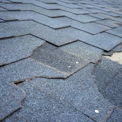 Close-up view of a worn asphalt shingle roof showing damaged and missing shingles. Nails are visible, indicating areas needing repair. The surface shows signs of weathering and age, highlighting the need for maintenance.