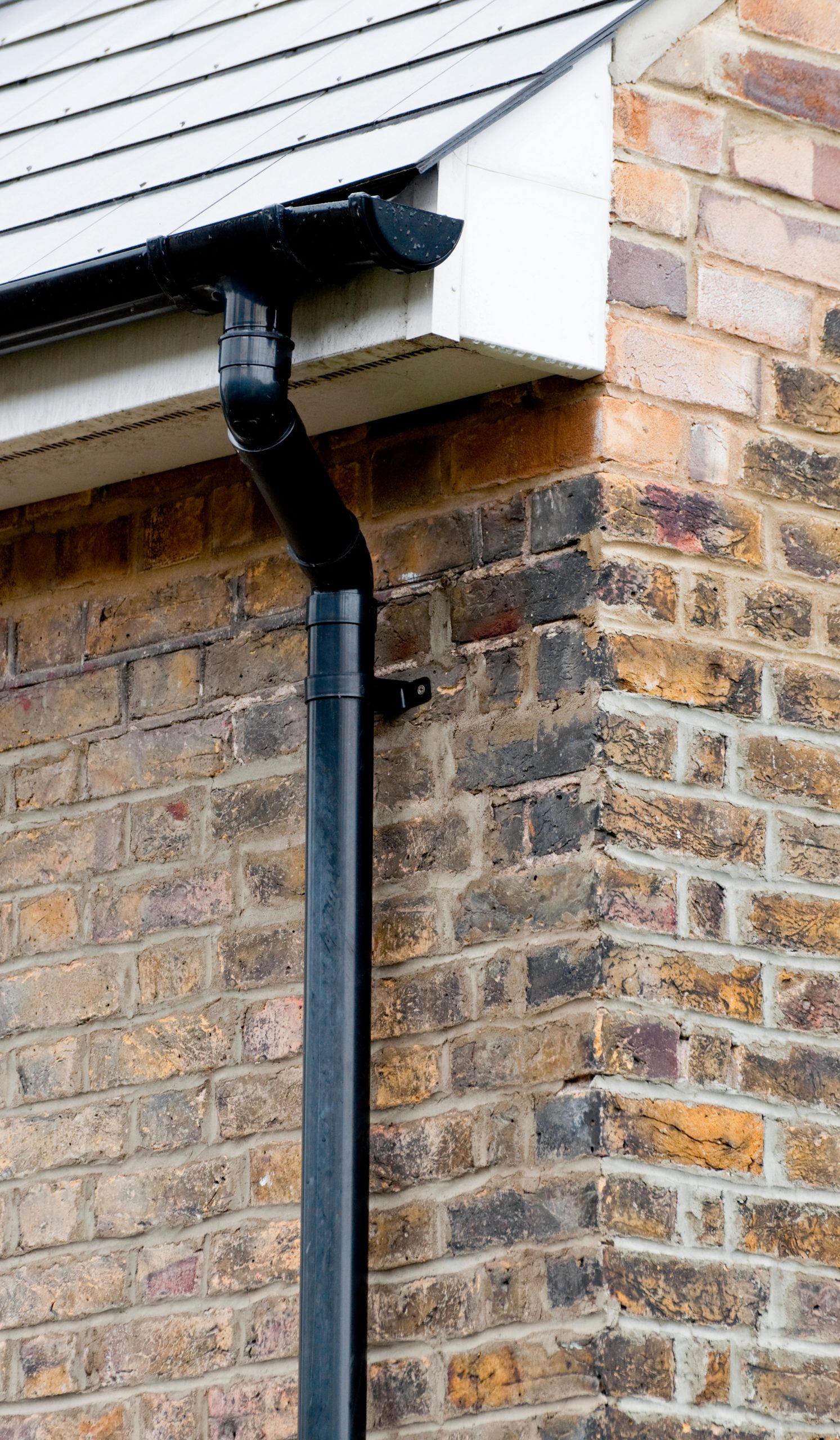 Close-up of a brick house corner with a black rain gutter and downspout attached near the roof. The bricks are of varying shades, adding texture and color contrast to the structure.
