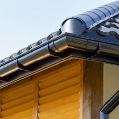 A close-up of a modern house roof with black metal gutters, neatly installed along the edge. The roof is covered with dark tiles, and wooden siding is visible beneath. The sky is clear in the background.