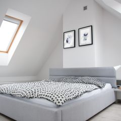 Minimalist bedroom with a modern gray bed, polka dot bedding, and a wooden bedside lamp. Two framed pictures hang on a white wall. A skylight window on the sloped ceiling lets in natural light. Light wood flooring complements the serene atmosphere.