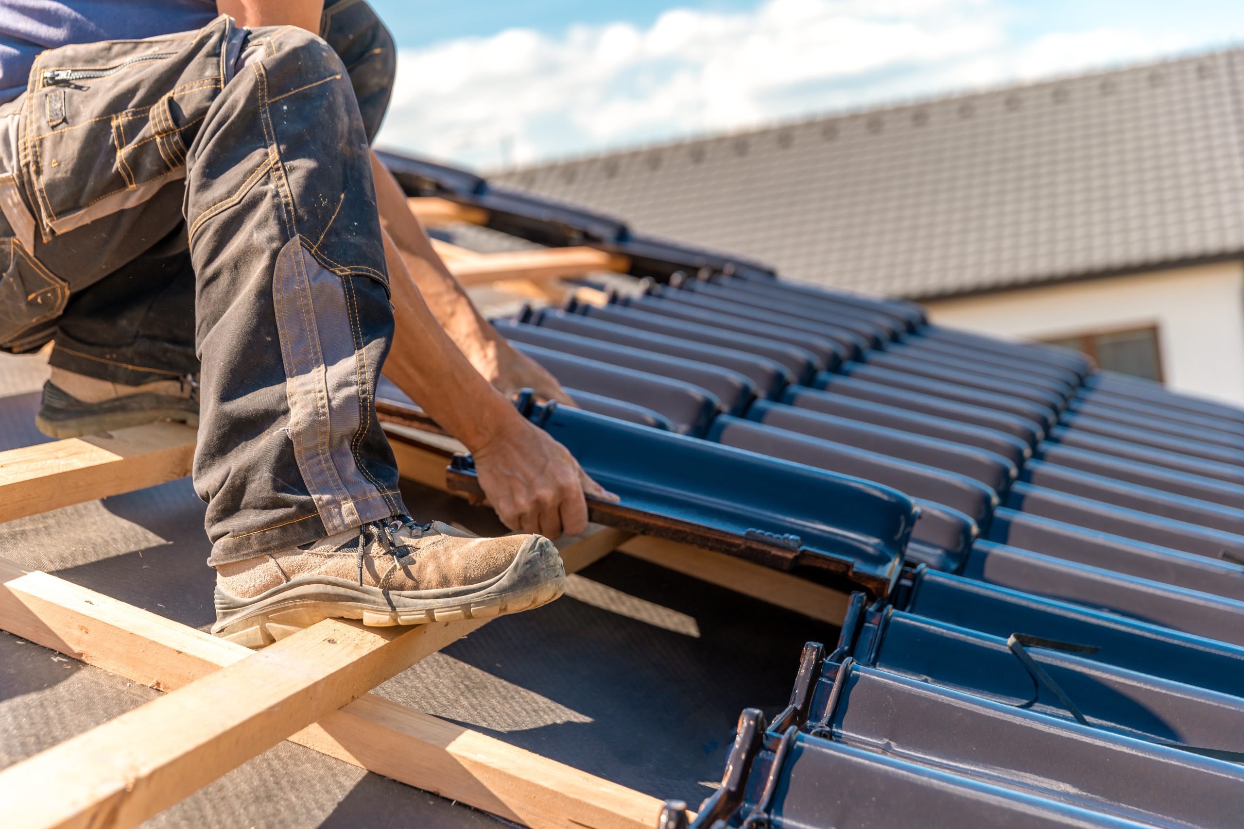 A construction worker installs black roof tiles on a pitched roof. The person is wearing work boots and jeans, kneeling on wooden beams. A house with a completed roof is visible in the background under a blue sky.