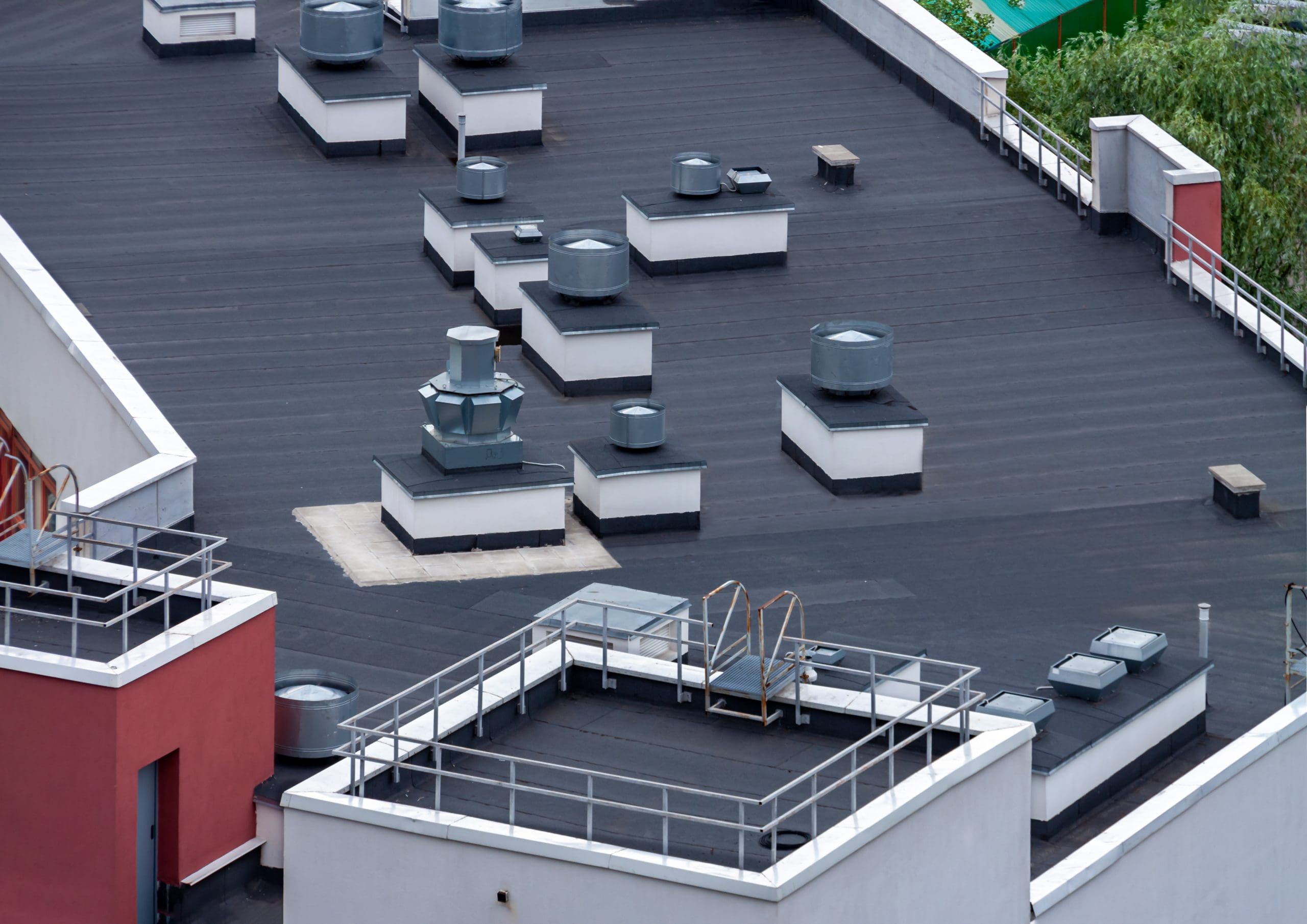 A rooftop with a black surface features several rectangular and cylindrical ventilation units. White and red building walls border the area, and a metal ladder is visible on one section. Green trees can be seen in the background.