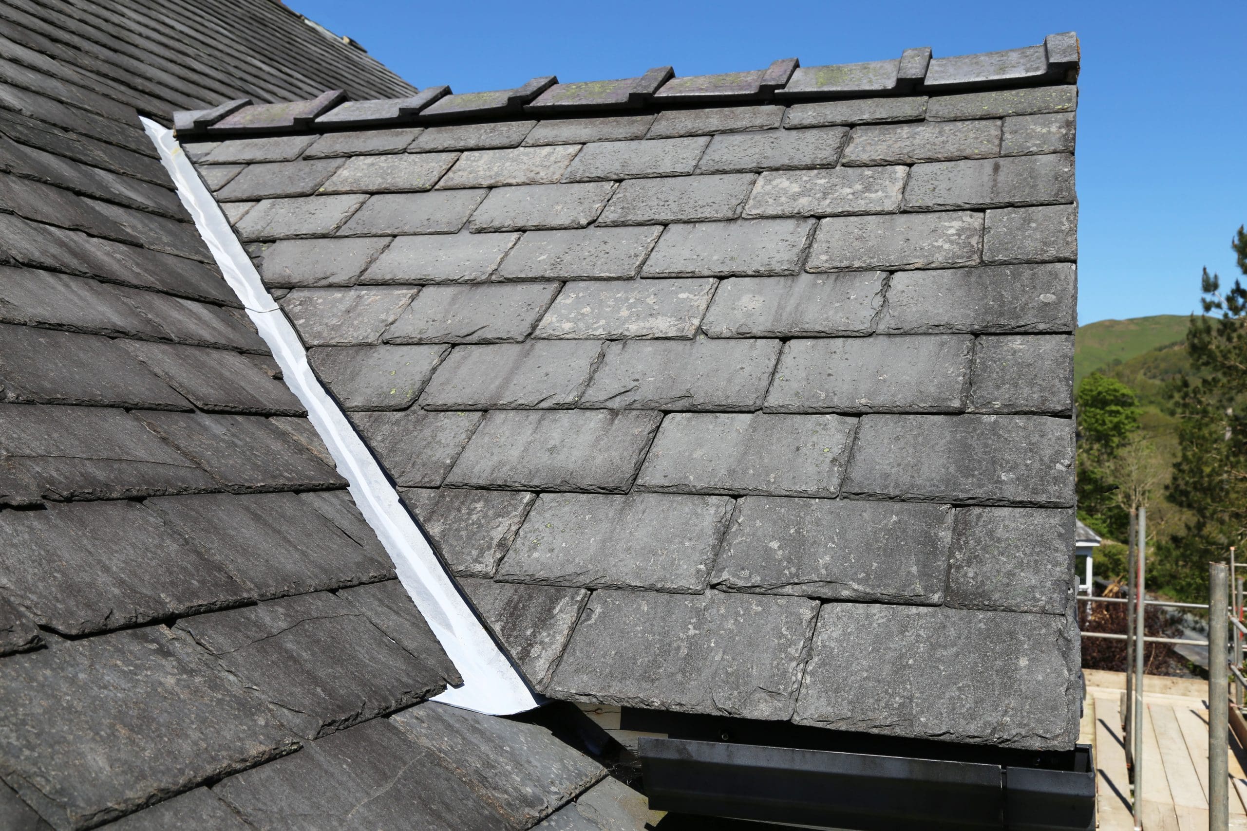 A close-up view of a slate roof with a ridge and flashing. The roofing tiles are arranged in overlapping rows, and the sky is clear and blue in the background. A few trees are visible at the edge of the roof.