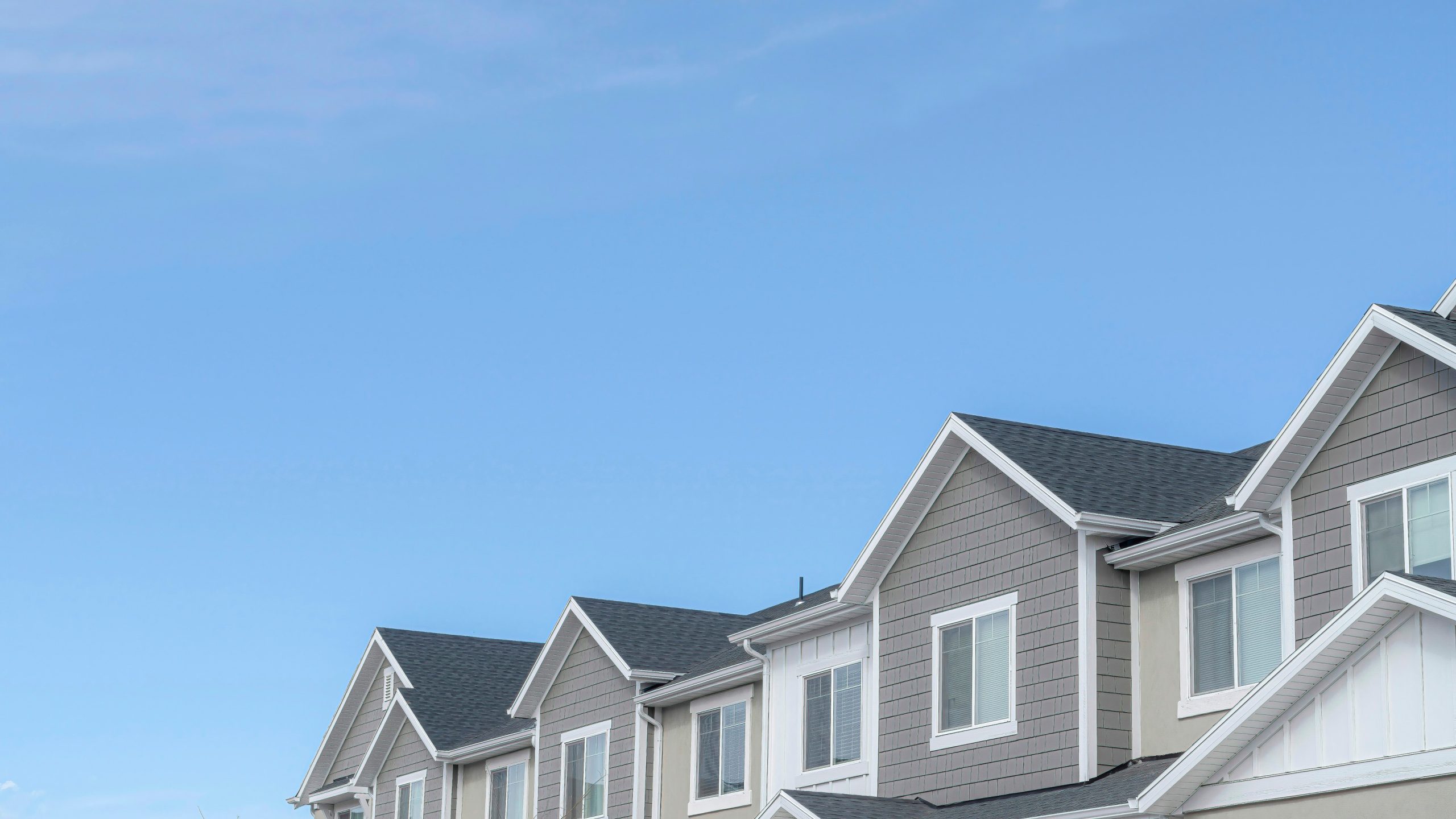 Row of modern townhouses with gray siding and white trim under a clear blue sky. The peaked roofs form a repeating pattern across the image.
