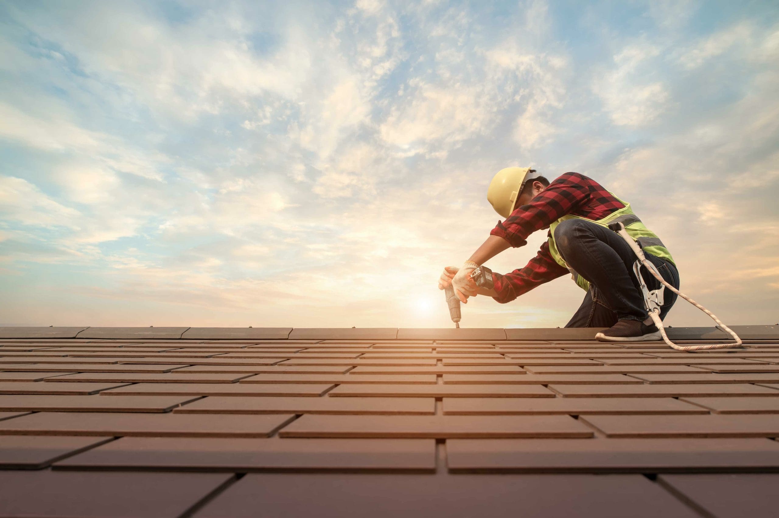 A construction worker wearing a hard hat and safety gear kneels while using a power drill on a rooftop against a backdrop of a cloudy sky with the sun setting on the horizon. A safety harness is visible beside the worker.