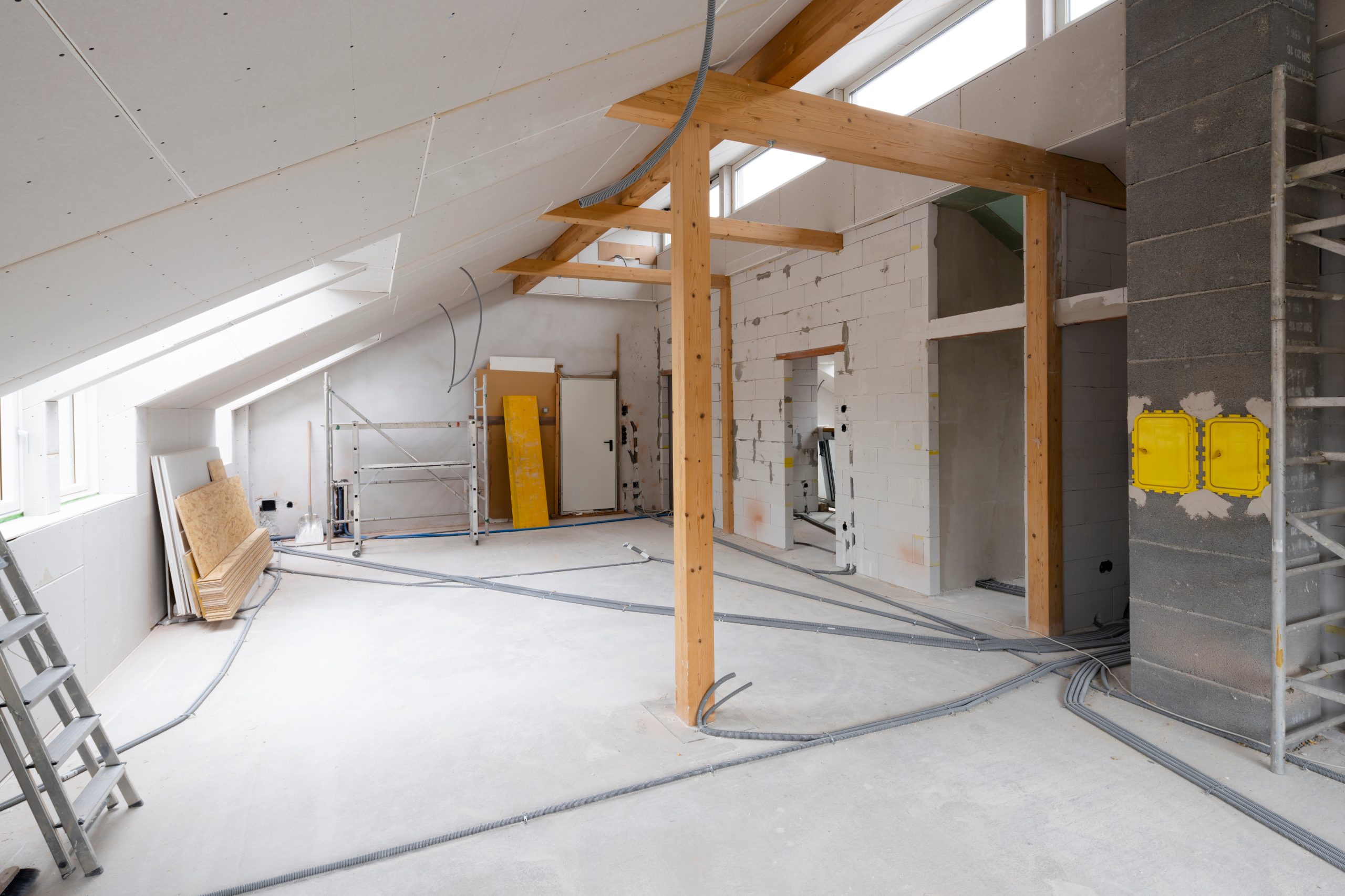 An unfinished attic under renovation, featuring exposed wooden beams, partially constructed walls, scattered electrical wiring, and construction scaffolding. Sunlight filters through windows, creating a bright workspace.