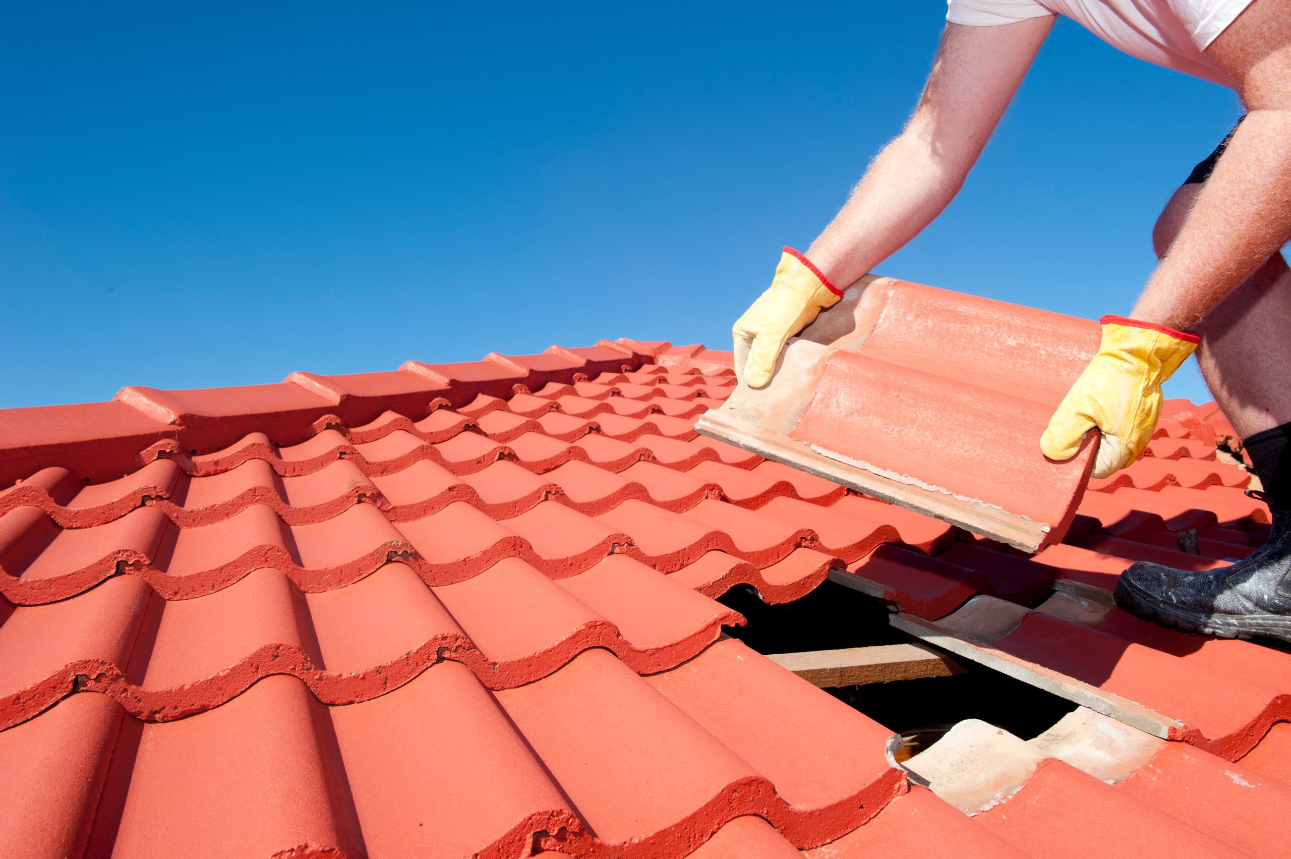 A person wearing gloves installs a red roof tile on a sunny day.