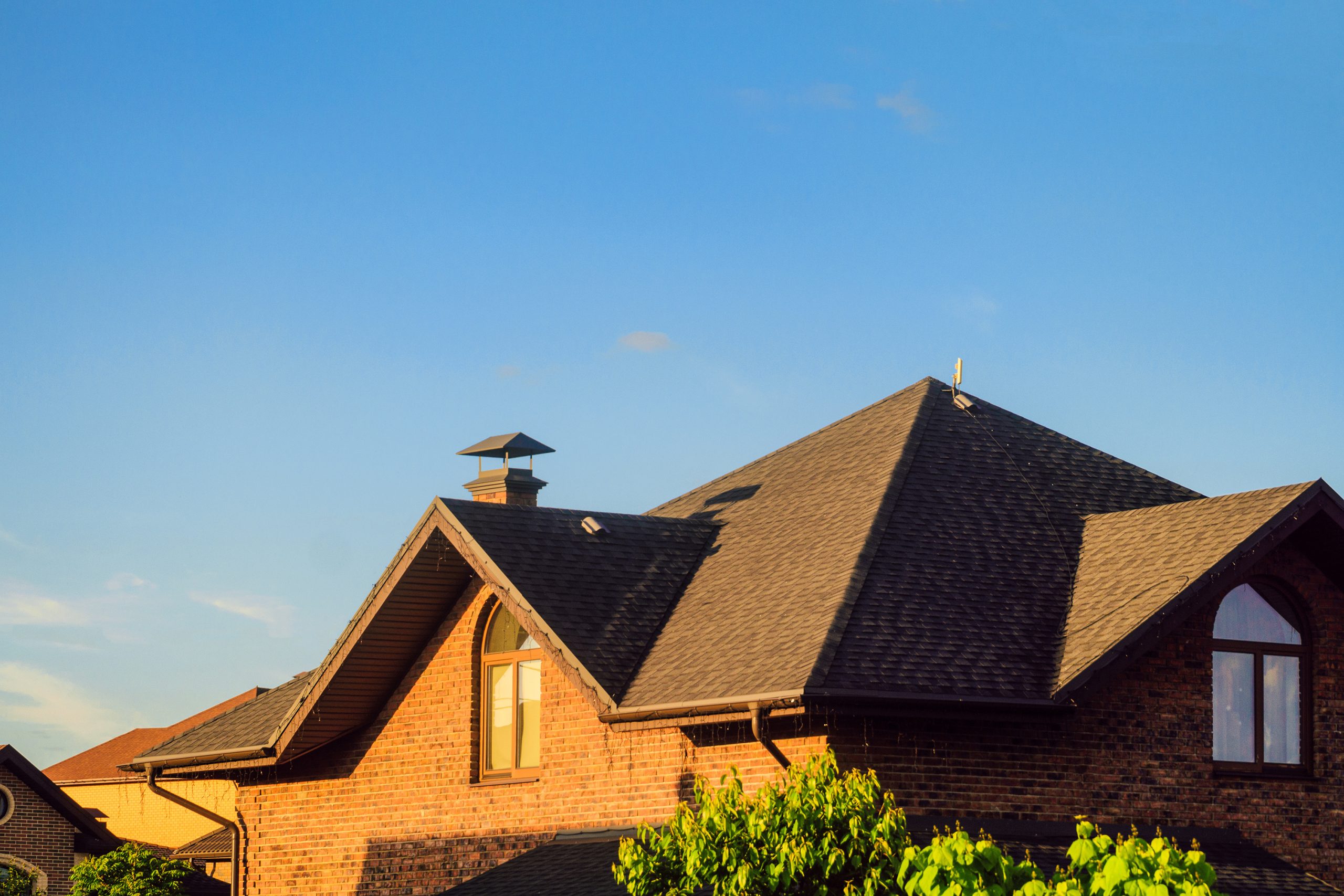 A red brick house with a large, steep, dark shingled roof and multiple dormer windows is shown. A chimney is visible against a clear blue sky, and green foliage is in the foreground.