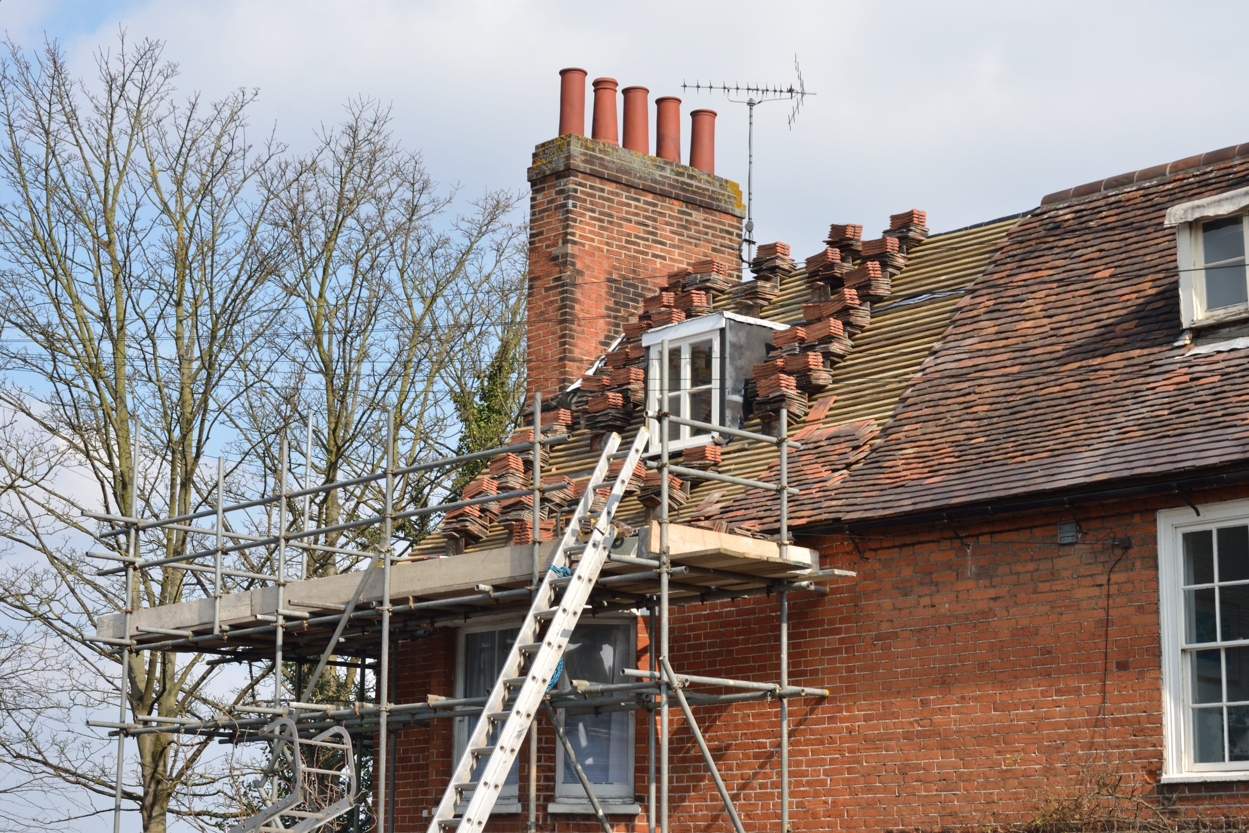 A brick house with a scaffold and ladder set up for roof repairs. Several tiles are missing near a dormer window. Leafless trees are visible in the background under a cloudy sky.