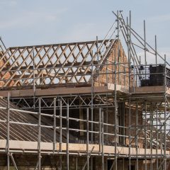 A partially constructed brick building with exposed wooden roof trusses is surrounded by metal scaffolding. A platform holds construction materials. The sky is clear, indicating daytime.