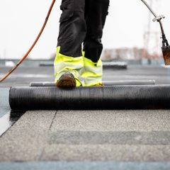 A worker in bright yellow safety gear applies a new layer of asphalt on a flat roof with a torch. The focus is on the worker's legs as they unroll the material. The area surrounds a cloudy sky backdrop.