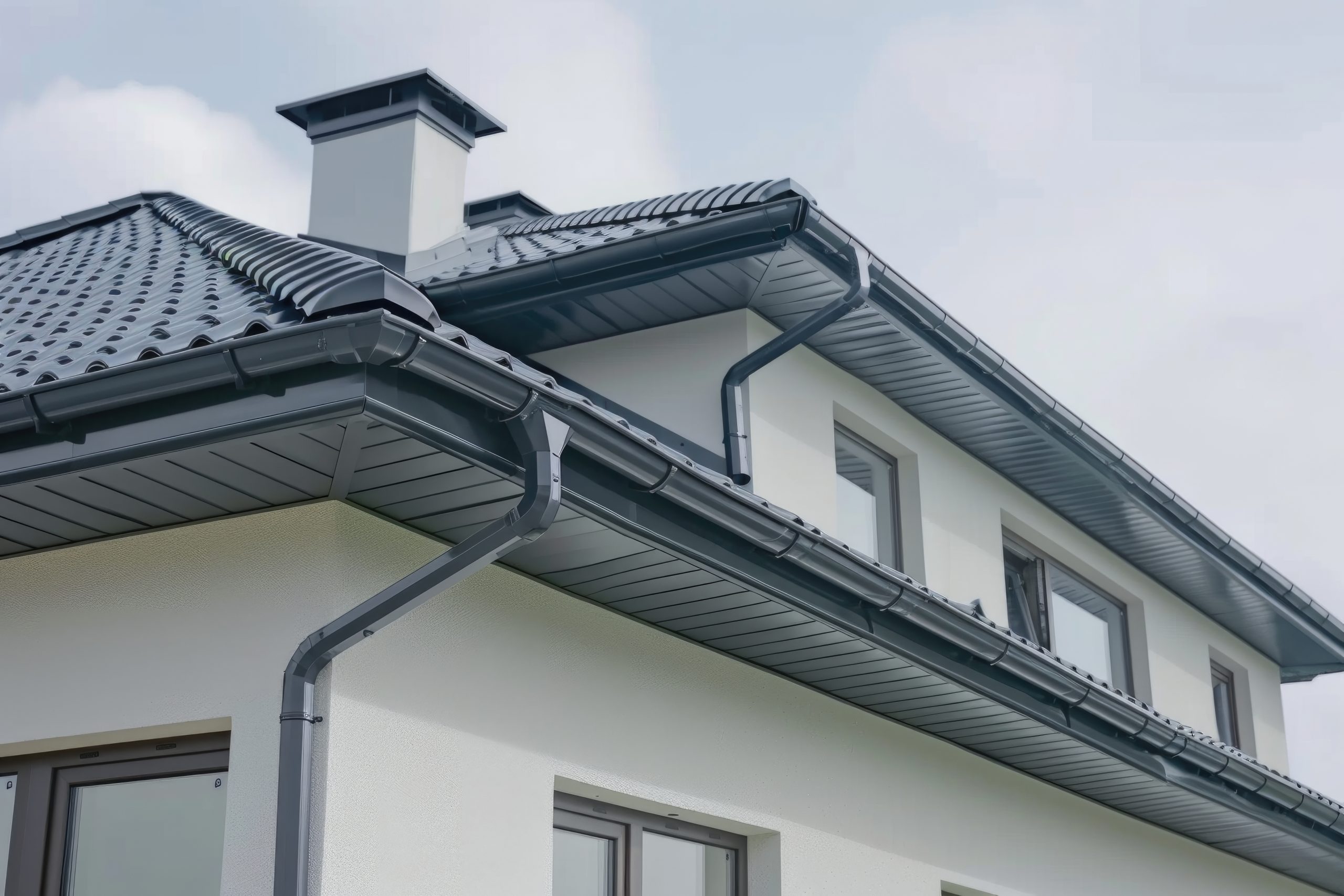 A modern white house with a sloped dark gray roof and matching gutters. The building features large windows and a chimney. The sky is overcast, adding a soft, muted backdrop.