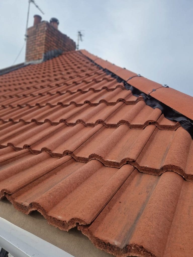 Close-up view of red terracotta roof tiles on a sloped roof, with a brick chimney and two TV antennas visible at the top against a cloudy sky.
