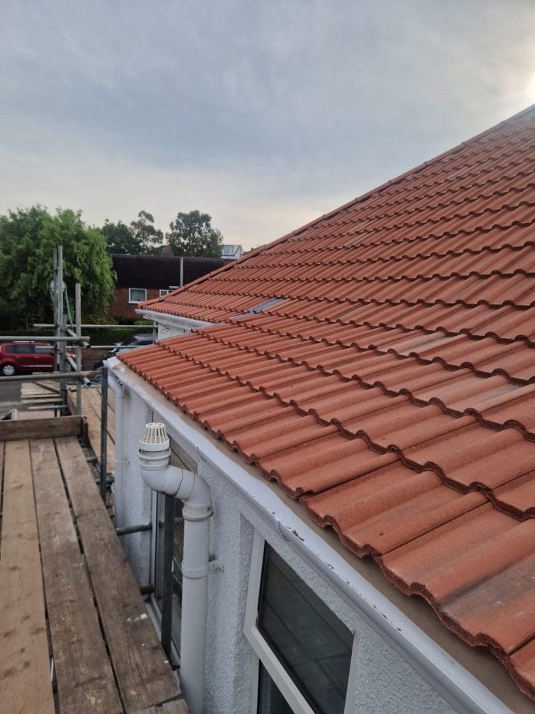 A view of a red tiled roof on a white house, taken from a scaffold platform. A drainpipe runs down the side, and trees, a red car, and other buildings are visible in the background under a cloudy sky.
