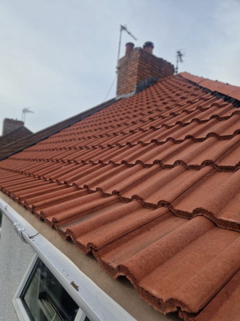 Close-up view of a red tiled roof with a chimney in the background on a cloudy day. The roof tiles are neatly arranged in rows, and part of a white wall and window are visible at the bottom left.