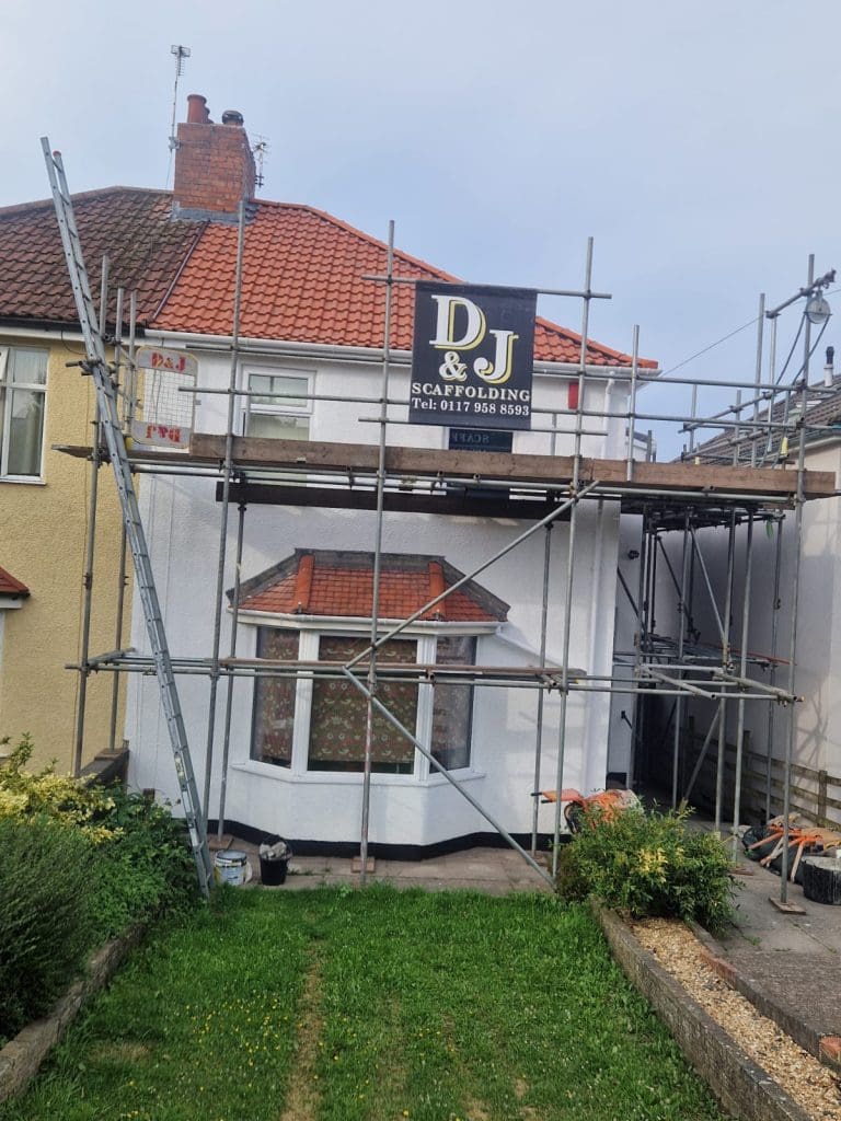 A house with red roof tiles and white walls is covered in scaffolding. A sign for "D&J Scaffolding" with a phone number is displayed at the top. The garden in front has a lawn and pathway.