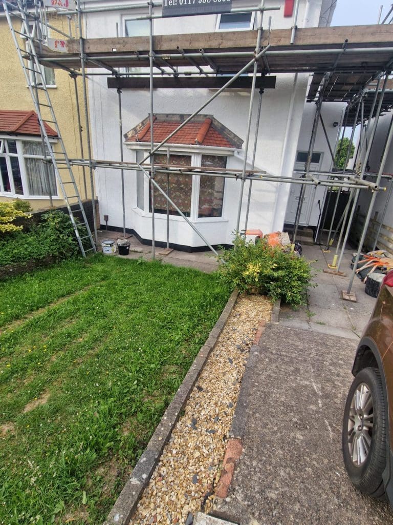 A house with scaffolding set up around the front, a bay window, and a grassy yard with a narrow gravel path leading to a driveway where a brown vehicle is parked. Various tools and buckets are on the ground.