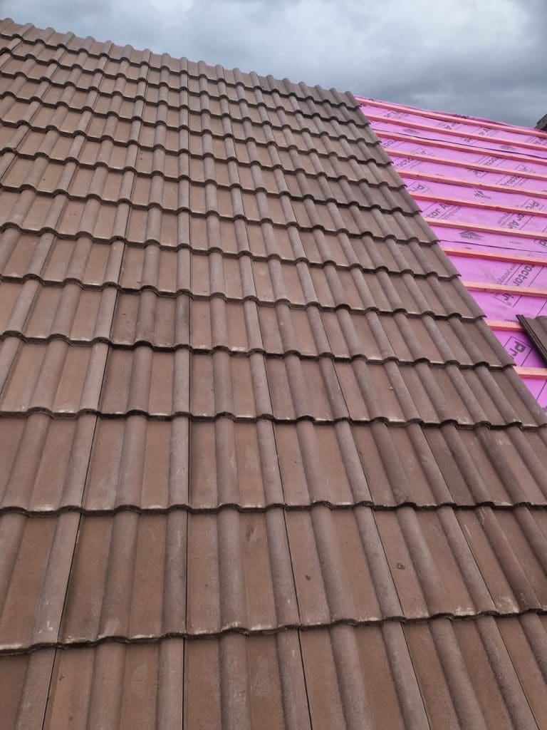 Brown roof tiles are installed on the left side of a sloped roof, while the right side shows a section covered with pink waterproof underlayment, awaiting tile installation under a cloudy sky.
