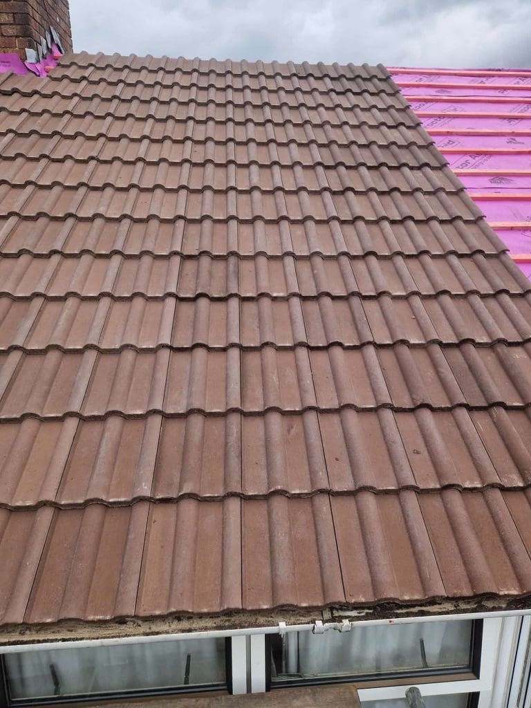 The image shows a roof partly covered with brown tiles, while the adjacent section is unfinished and covered with bright pink waterproof roofing underlayment. A cloudy sky is visible above.