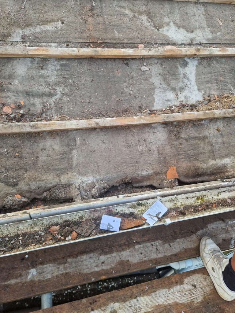 A close-up of a weathered, dirty wooden surface with several planks, scattered debris, and two pieces of paper. The lower right corner shows part of a white sneaker.