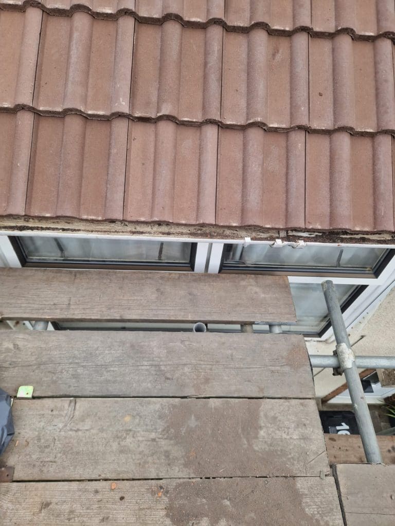A view from above of a tiled roof, white window frames below it, and wooden scaffolding planks with metal supports in the foreground.