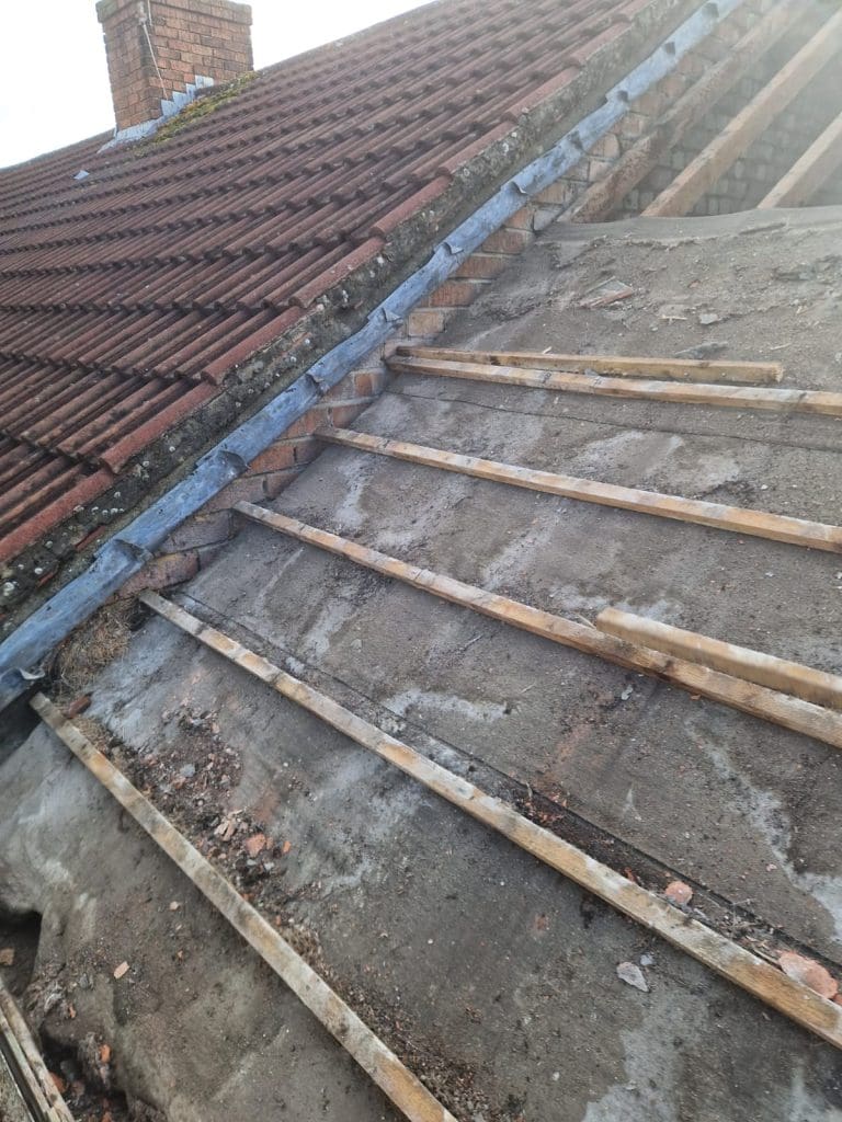 Part of a tiled roof under repair, with some tiles removed to reveal wooden battens and the underlying concrete surface; a brick chimney is visible in the background.