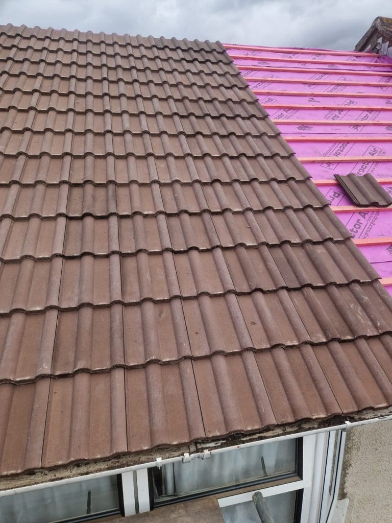 A roof under construction with brown roof tiles partially installed. The right side shows exposed bright pink waterproof underlayment, while the left side is covered with tiles. A cloudy sky is visible in the background.