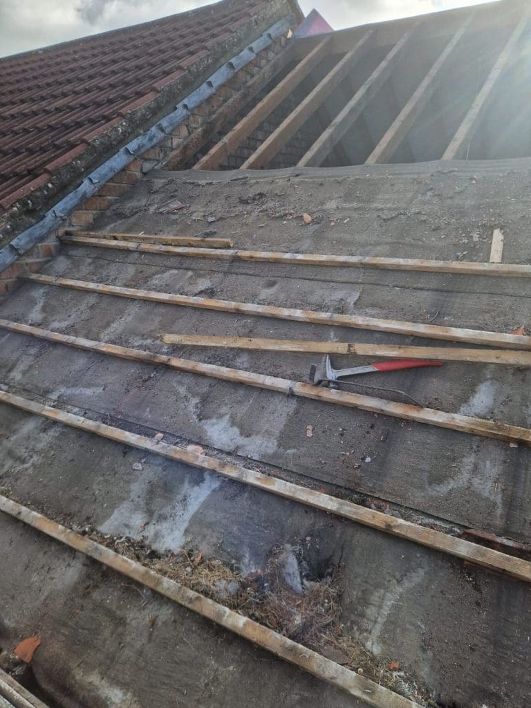 A partially stripped roof shows exposed wooden beams and battens, with a hammer and crowbar lying on the concrete surface beneath the removed tiles. The sky is cloudy in the background.