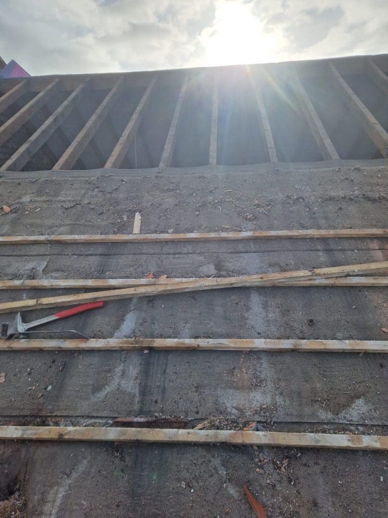 Wooden planks and construction tools rest on a concrete surface in sunlight, with wooden beams and a bright sky in the background, suggesting an ongoing building project.