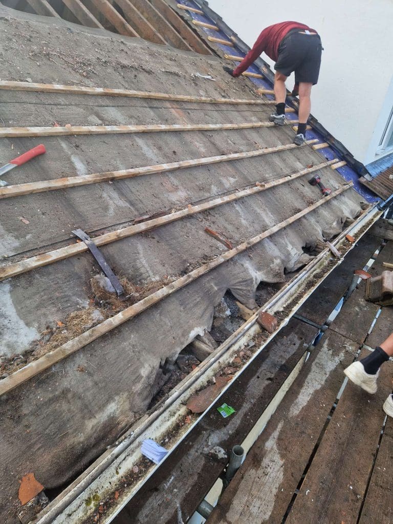 A person in shorts and a red sweater works on a sloped roof stripped of tiles, exposing wooden battens and underlying materials, while another person's legs are visible at the edge of scaffolding. Tools and debris are scattered around.