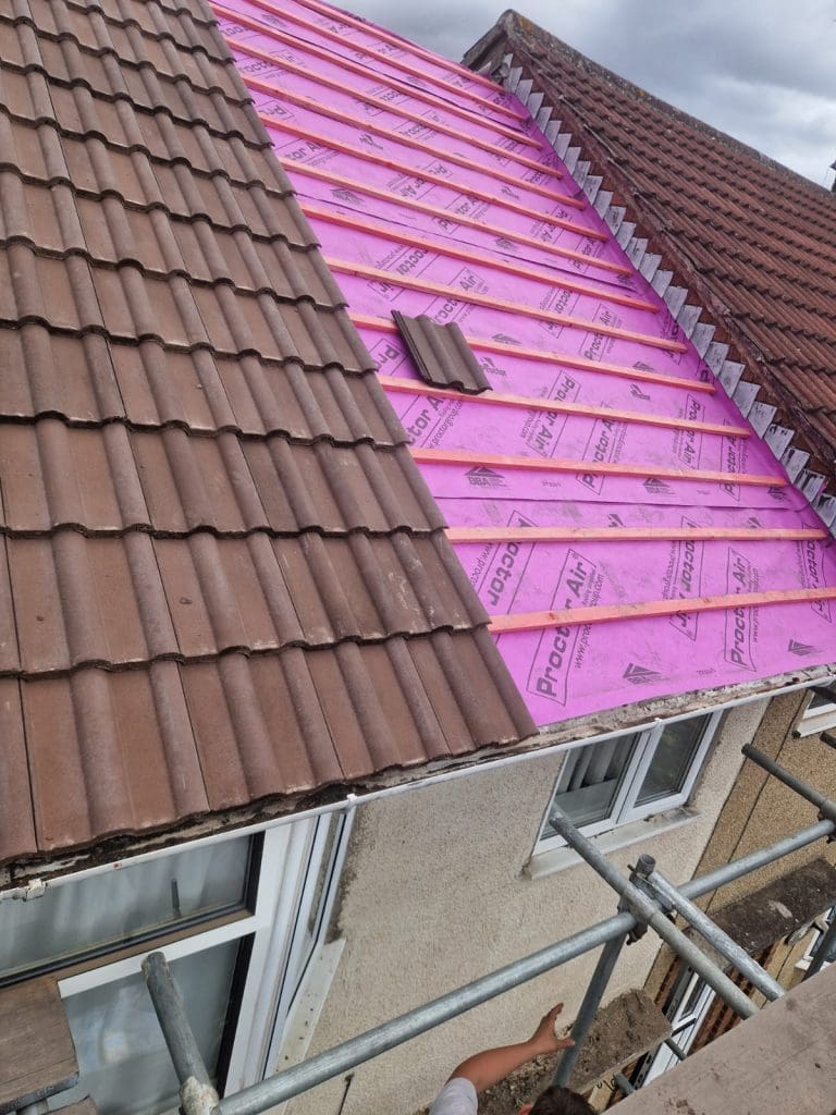 A sloped house roof partly covered in brown tiles, with the other half showing bright pink waterproof underlayment and no tiles yet. Scaffolding and part of a person's arm are visible at the bottom.