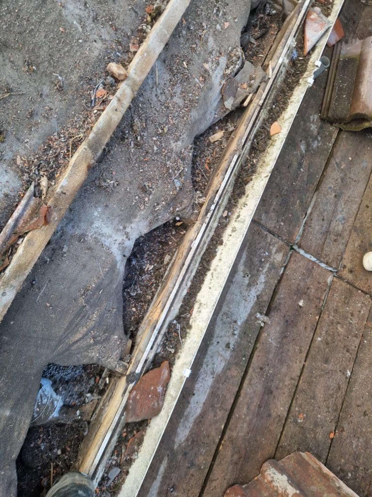 A close-up view of a damaged roof showing torn underlay, exposed wooden beams, scattered debris, and broken roof tiles on a wooden surface.
