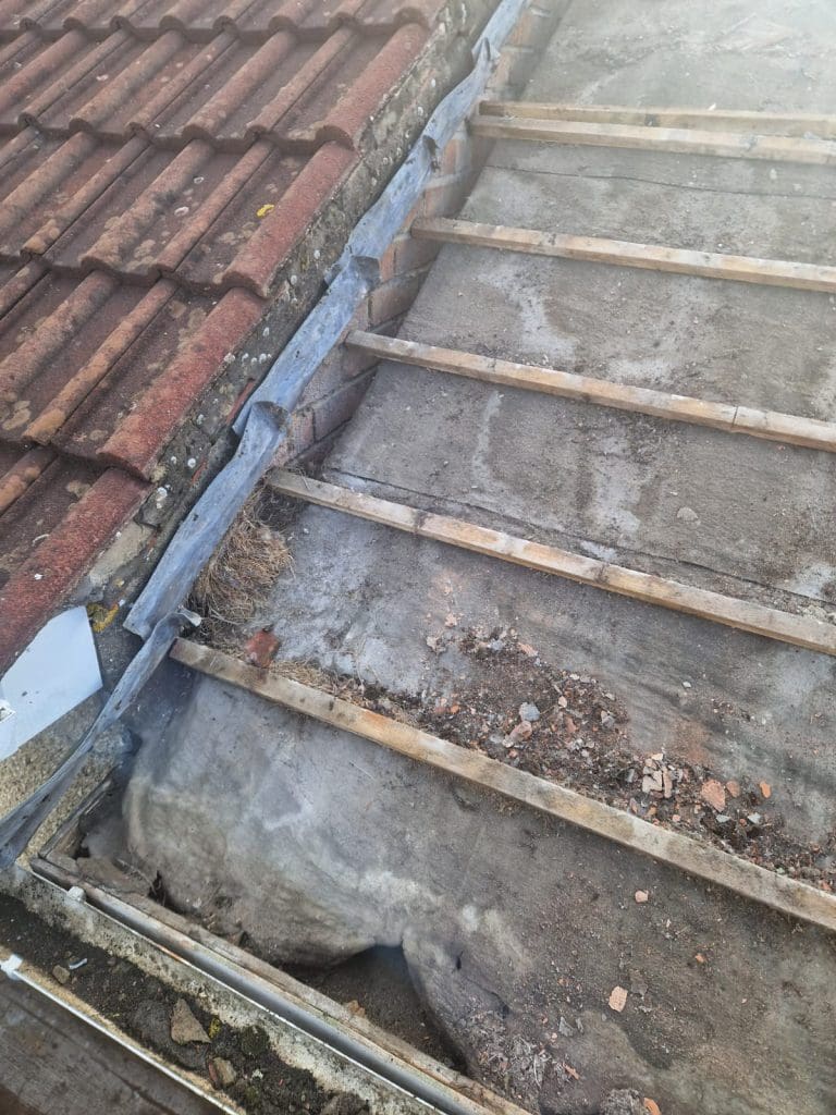 A roof under repair with old red tiles partially removed, exposing wooden beams and underlying surface. Debris and dust are visible on the exposed area.
