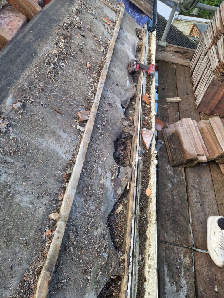 A damaged, debris-covered roof with broken tiles and exposed wood beams. A red drill sits on the roof, and stacked tiles and construction materials are nearby. Part of a boot is visible in the lower right corner.