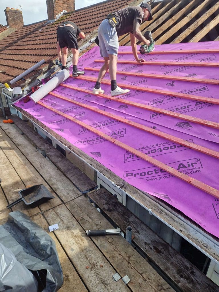 Two workers install wooden battens over a bright pink roofing membrane labeled "Proctor Air" on a partially tiled roof. Tools and materials are scattered on the wooden scaffolding platform below.
