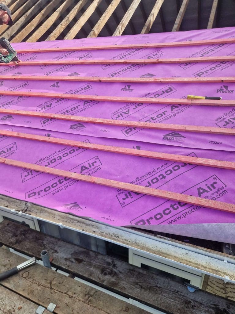 A roof under construction with pink Proctor Air membrane and wooden battens installed across it; roof rafters are visible at the top and a worker's gloved hand appears on the left.