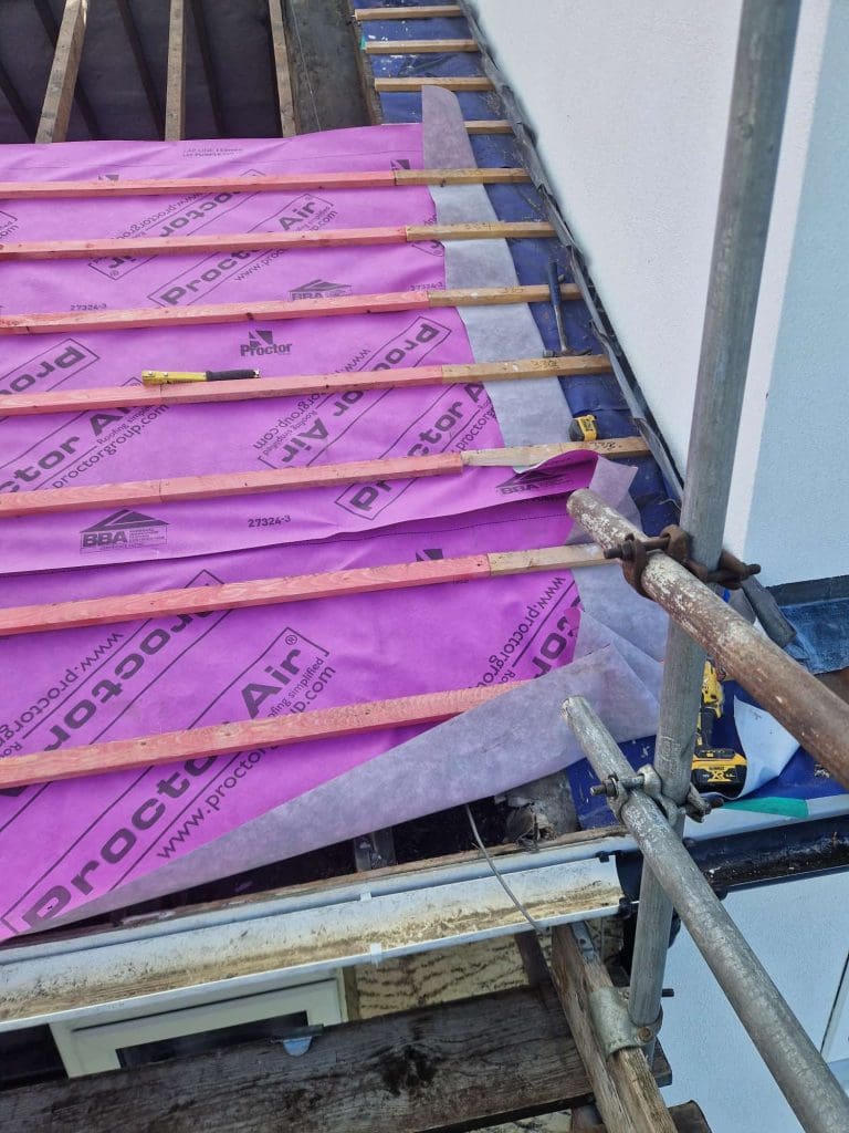 Roof under construction with wooden battens and purple Proctor Air membrane partially installed, scaffolding in place, tools visible on the membrane, and a white wall on the right.